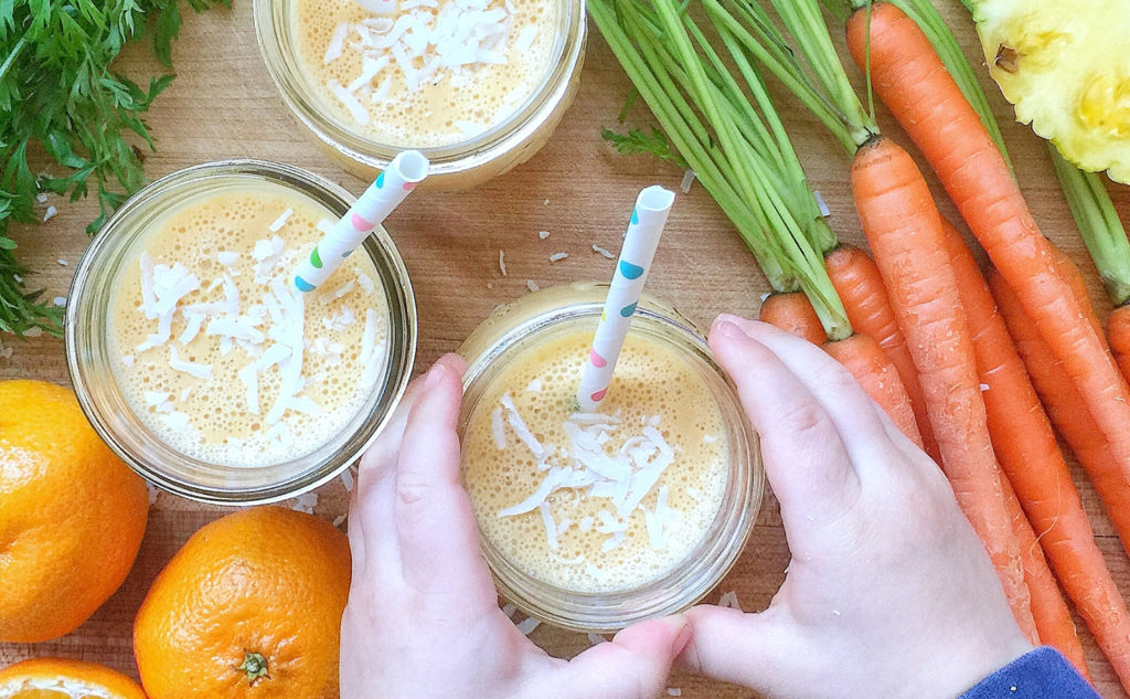 A small child's hands picking up a jar filled with a smoothie and a straw. There are 2 other smoothie-filled jars on the counter and are surrounded by oranges, carrots, and fresh herbs. This recipe is included in the list of best smoothie recipes for kids.