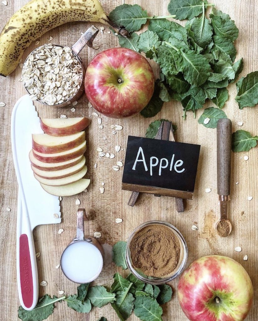 A wooden surface topped with a plastic, kids' chef knife, slices of apple, 2 whole apples, fresh greens, a banana, a measuring cup full of oats, a measurng cup full of milk, a measuring spoon filled with cinnamon, a small jar filled with cinnamon, a chalkboard mini easel with the word "apple" on it. This recipe is included in the list of best smoothie recipes for kids.