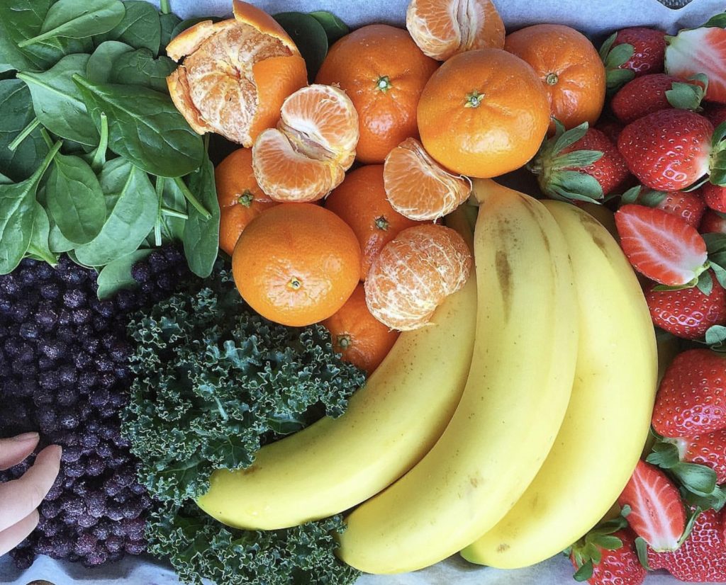 Bananas, clementines (whole and unpeeled as well as a few unpeeled and broken apart into segments), kale, strawberries, baby spinach leaves, and frozen blueberries all arranged on a countertop. A hand is picking up a blueberry. These are all ingredients suggested as options to make the smoothie recipes for kids.