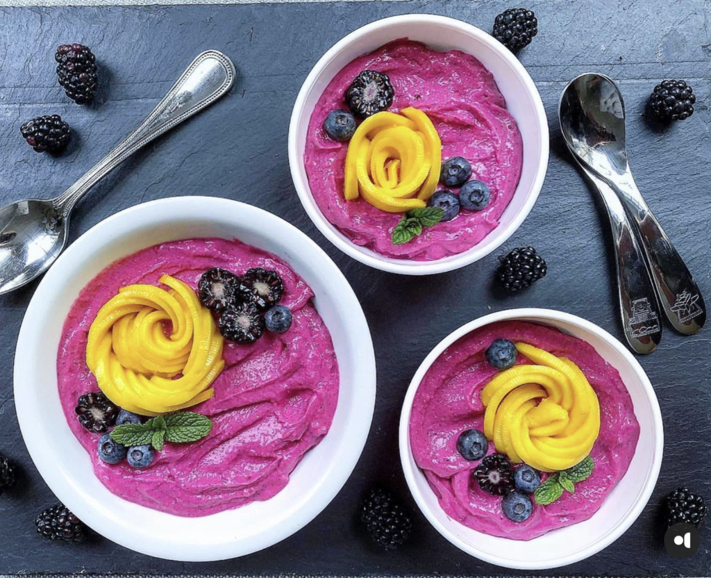 3 bowls filled with purple dragonfruit smoothie and all topped with fresh berries and mango. Bowls are sitting on a counterop and surrounded by a few spoons and fresh blackberries strewn about the countertop. This recipe is included in the list of best smoothie recipes for kids.