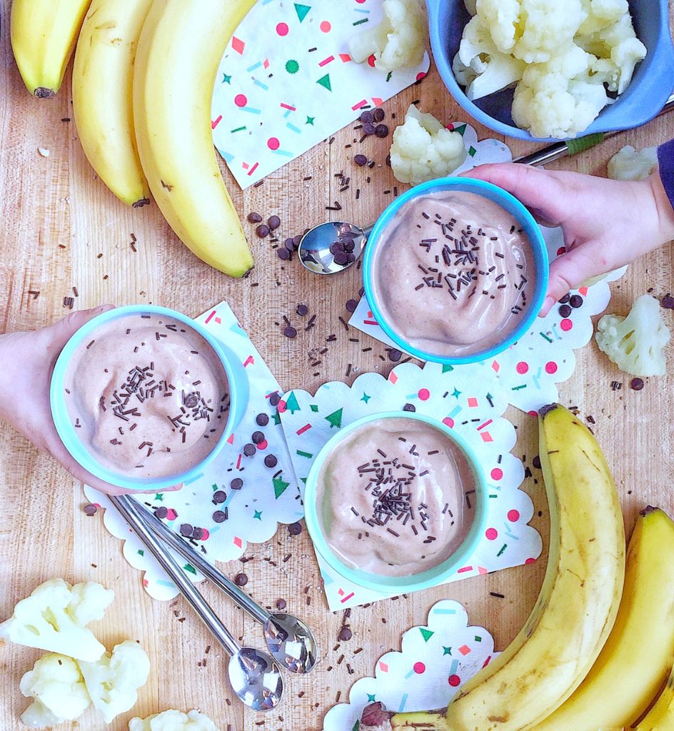 2 smoothies with sprinkles being picked up from a wooden countertop. A third smoothie with sprinkles is sitting on a napkin on the countertop and is surrounded by several bananas, napkins, and cauliflower in a bowl. Pieces of cauliflower, sprinkles, and mini chocolate chips are strewn about the countertop. This recipe is included in the list of best smoothie recipes for kids.