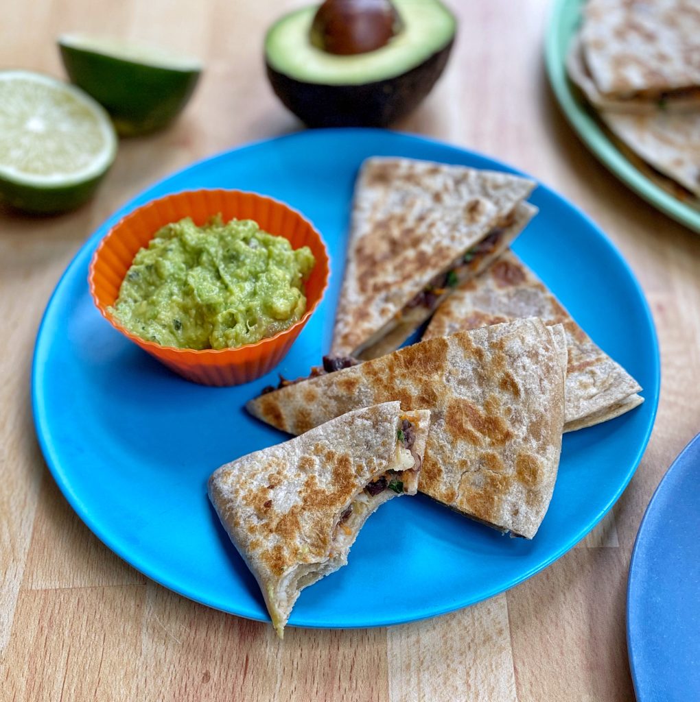 Quesadillas cut into triangles and a side of guacamole sitting on a blue plate. The plate issitting on a  wooden countertop and surrounded by sliced limes, a halved avocado, and another plate filled with quesadilla points, 