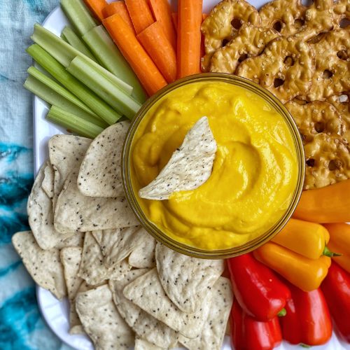 Cheese dip made from cauliflower and carrots in a bowl with a chip dipping in. Surrounded by colorful veggies, more chips, and pretzels.