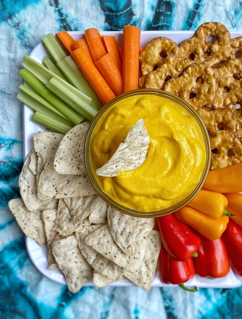 Cheese dip made from cauliflower and carrots in a bowl with a chip dipping in. Surrounded by colorful veggies, more chips, and pretzels.