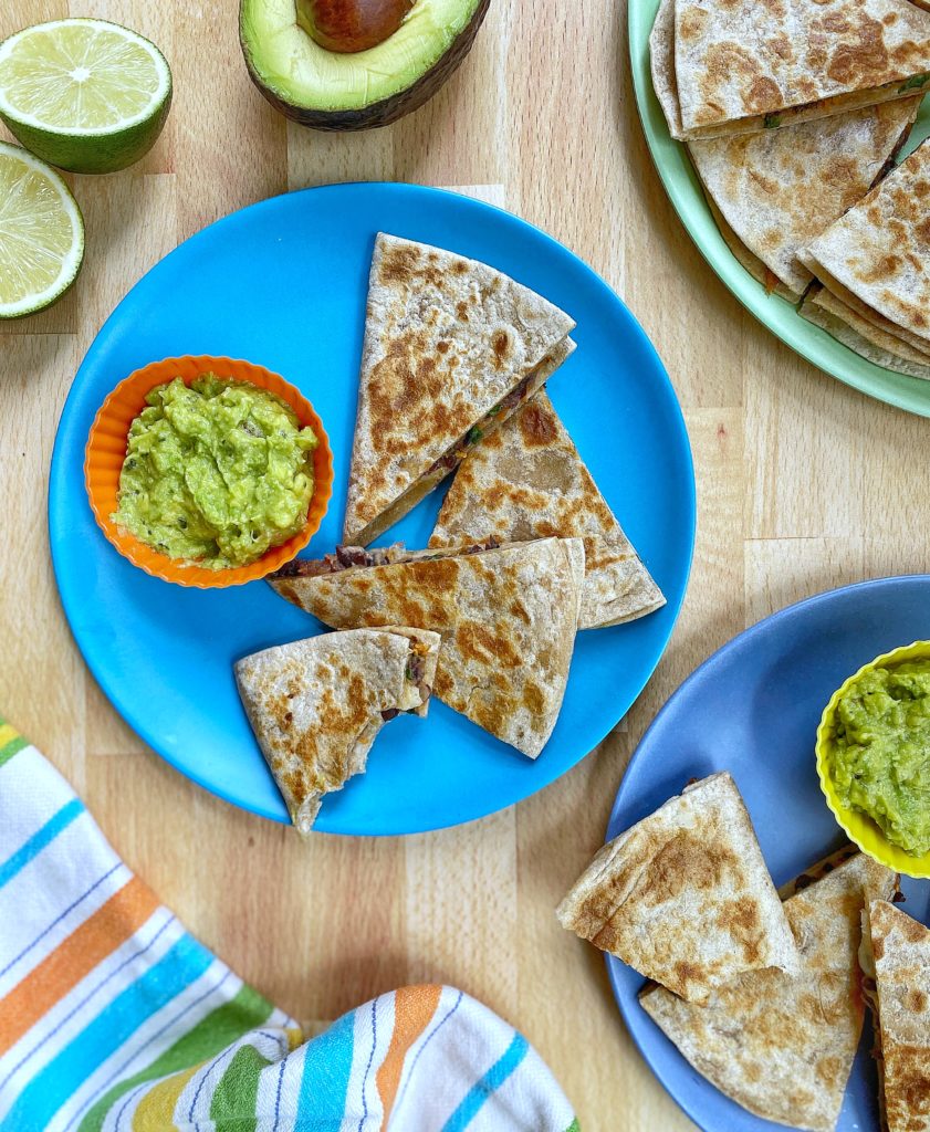 Quesadillas cut into triangles and a side of guacamole sitting on two blue plates. Plates are sitting on  a  wooden countertop and surrounded by a napkin, sliced limes, a halved avocado, and another plate filled with quesadilla points, 