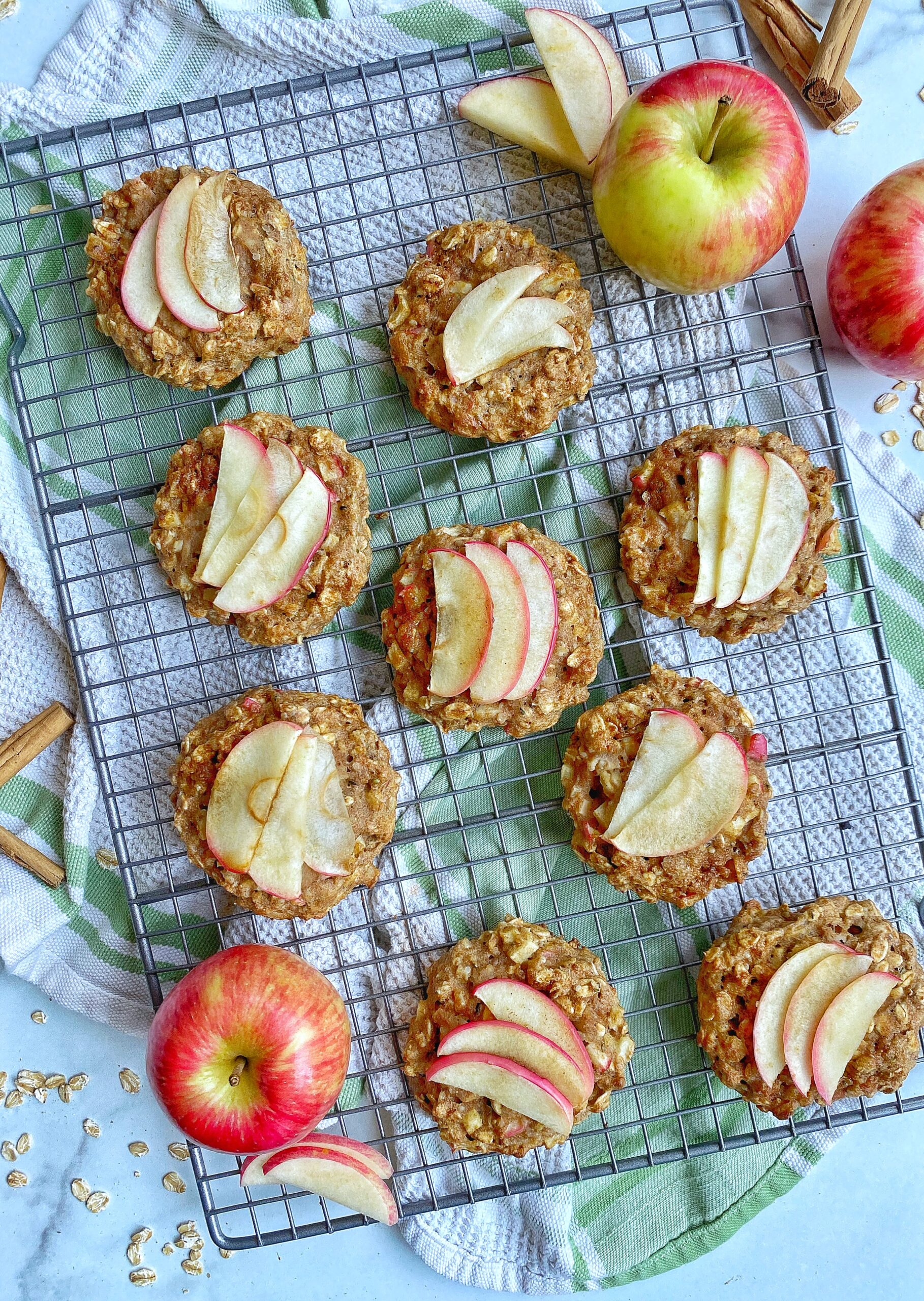 Breakfast cookies topped with apple slices sitting on top of a wire rack on top of a kitchen towel. Whole apples, apple slices, cinnamon sticks, and spilled oats are surrounding the breakfast cookies.