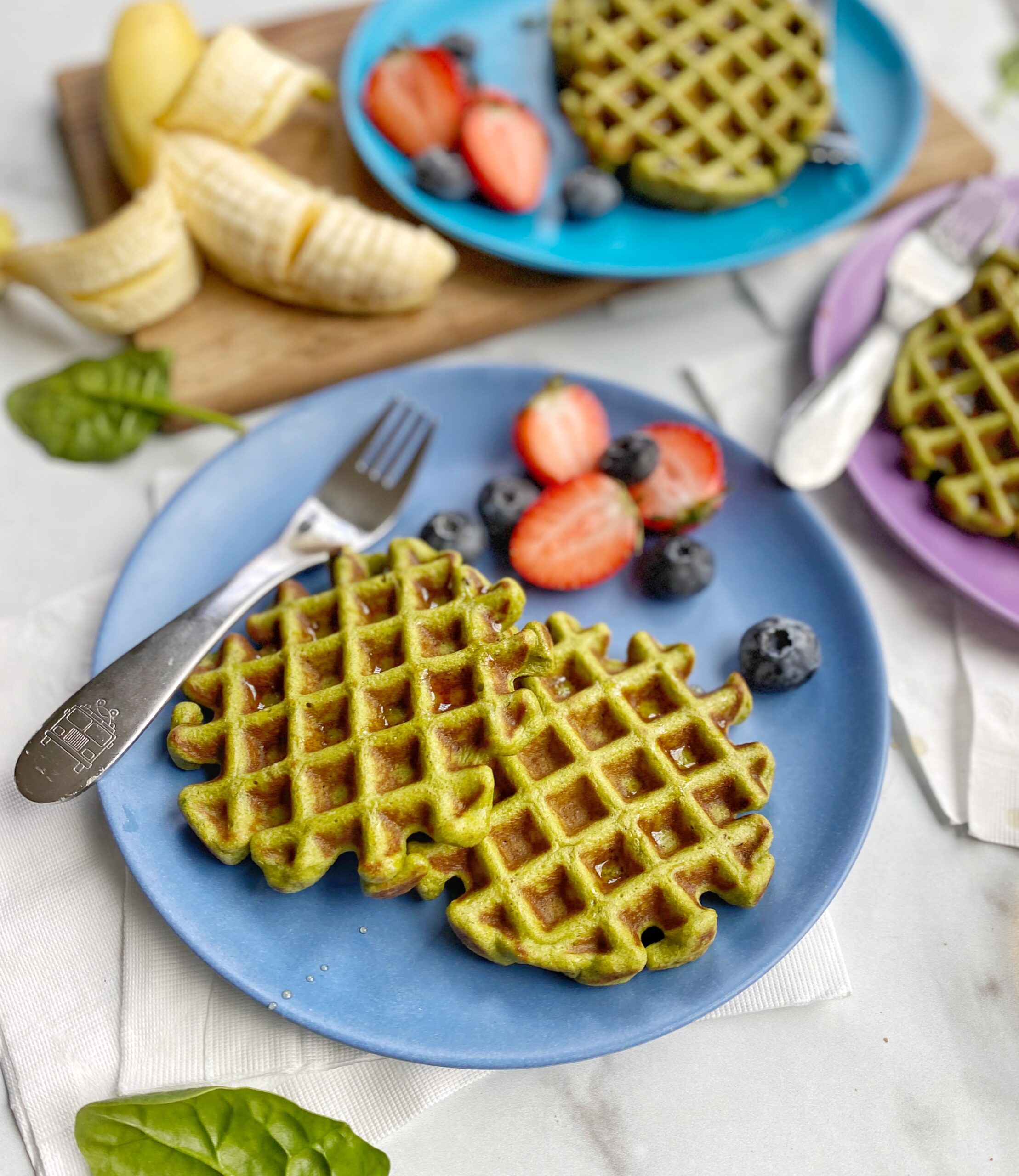 3 plates with Spinach Banana Waffles with strawberries and blueberries also on the plate. A sliced banana still half in the peel on a cutting board surrounding the plates on a counter. Counter has pieces of spinach strewn about the counter and a bowl of syrup with a spoon on the counter.