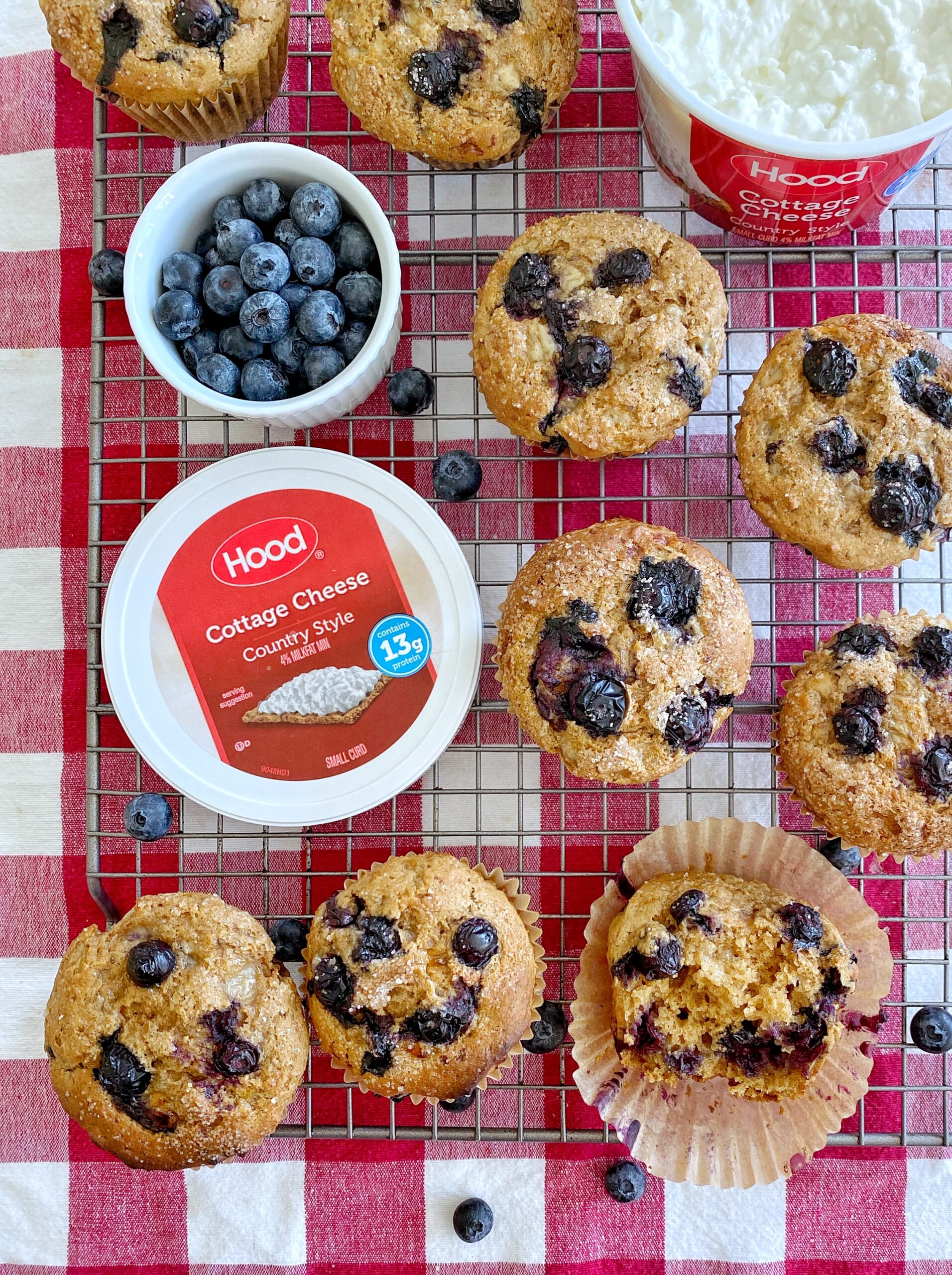 Blueberry muffins in paper liners on a wire rack sitting atop a red and white checkered tablecloth. A ramekin of blueberries and an open container of cottage cheese are mixed in with the muffins.