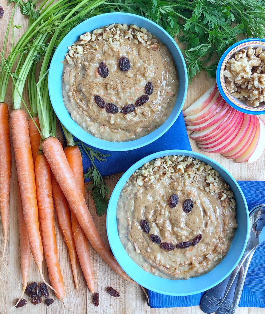 Two bowls of oatmeal sitting on a table surrounded by whole carrots, raisins strewn about the countertop, slices, of apples, a small bowl of walnuts, and a few spoons and napkins. Raisins are arranged in the shape of faces on the bowls of oatmeal.