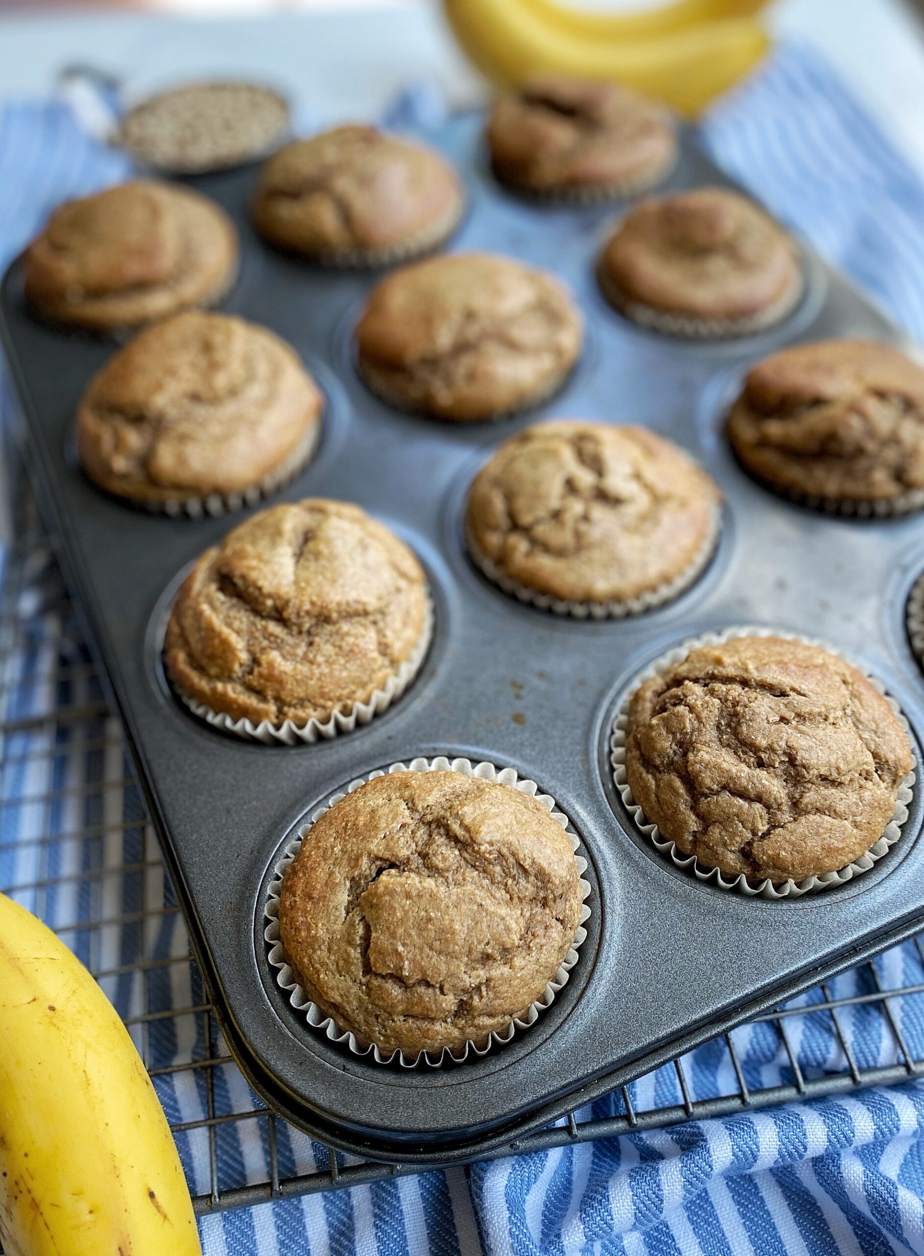Baked muffins in a cupcake tin filled with paper muffin liners. Muffin tin is sitting on a cooling rack on top of a kitchen towel and is surrounded by bananas.