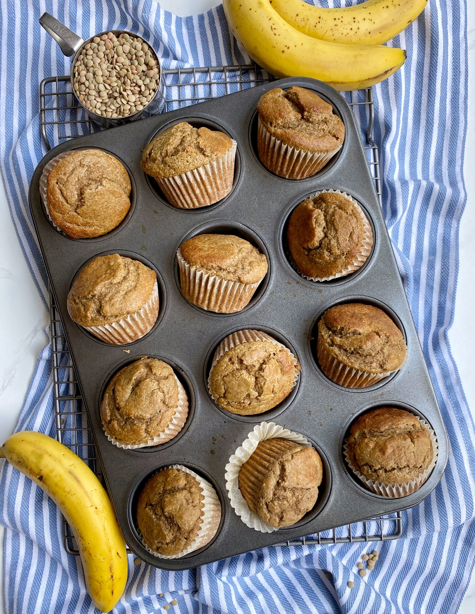 Baked muffins in a cupcake tin filled with paper muffin liners. Muffin tin is sitting on a cooling rack on top of a kitchen towel and is surrounded by bananas and a measuring cup full of lentils.