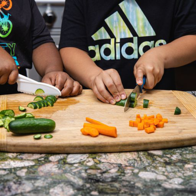 Two children chopping vegetables (carrots and cucumbers). One child is using a plastic kids' knife and the other is using a metal knife.