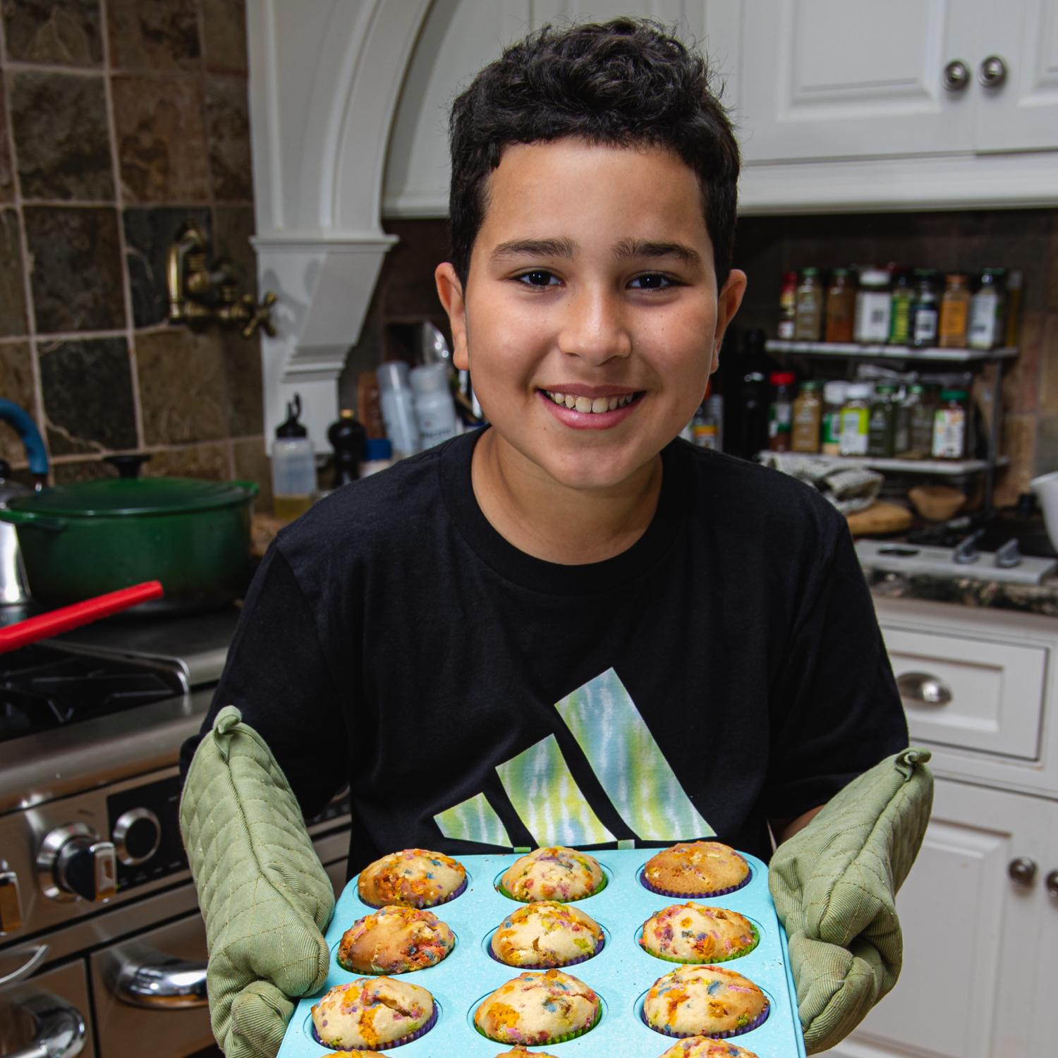 Boy in kitchen with oven mitts. He is holding a muffin tin filled with muffins.